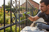 erstelle mir ein bild wo ein handwerker mit einem pinsel mit grünem handgriff, einen metallzaun im außenbereich mit anthrazitgrauem lack streicht